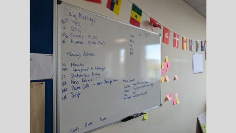 A wall-mounted white board with notes written in blue market pen including details of daily meetings and actions. Surrounding the board are international flags and sticky notes.