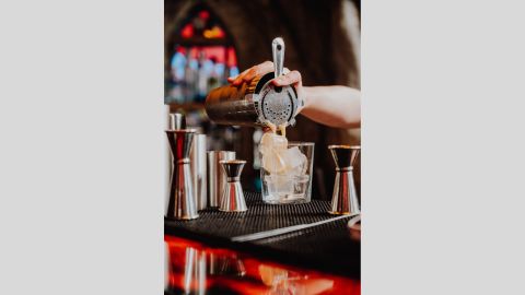 A cream coloured liquid poured from a cocktail shaker into a glass of ice. Measuring cups surround them.