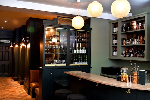 The bar at Virgin Mary Dublin consisting of a marble bar, sphere ceiling light globes, a large dark glass fronted cupboard with bottles and wall mounted open green units containing bottles. Snug seating and parquet flooring is visible in the background.