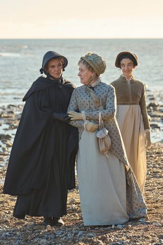 Episodic photo: on the pebby shore with the sea behind, Cassy and Mrs Austen comfort one another while Jane looks on. The are dressed in long dresses and coats of the Georgian and Regency era