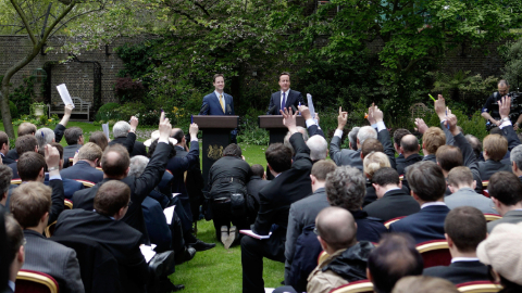 Nick Clegg and David Cameron, both white middle aged men in suits, stand at lecterns in front of trees while taking questions from dozens of journalist sat on chairs in the foreground.