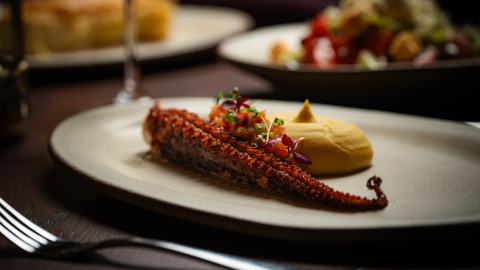 A low angled photo of grilled octopus on a plate with cutlery and other meals in the background