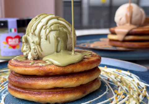 Three pancakes stacked on a plate with a scoop of green ice-cream bieng drizzled with a green liquid - there are other packages blurred in the background 