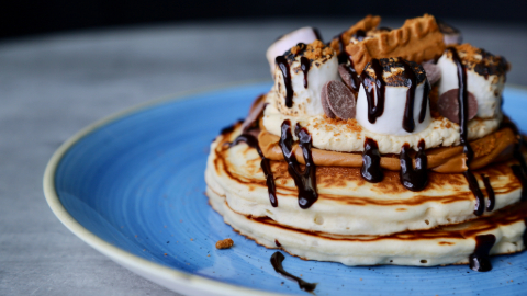 Two pancakes on a blue plate piled with peanut butter, marshmellows, broken Lotus Biscoff biscuits and drizzled with chocolate sauce