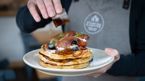 An unseen person wearing an apron holding a plate of three pancakes wiith bacon and fruit being drizzled with a dark syrup