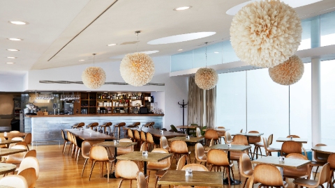 A light-filled restaurant interior with a floor to ceiling window, sloped white ceiling, wood floor, wooden table and chairs, five ceiling-hung cream pompom decorations and a bar in the background