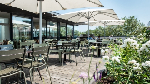 A restaurant roof terrace on a sunny day with green chairs, large white parasols, flower beds and a blue sky.