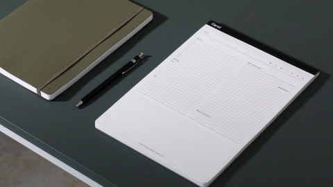 On a dark green table sits an olive green notebook with band, a black pen, and a white planner.