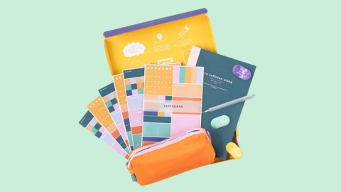 On a green pastel background, a birds eye view of an assortment of brightly coloured notebooks, planners, pencil case and other stationery