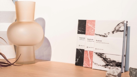 Three journals with black, pink and white marble effect covers leant against a wall on a wooden shelf. They are surrounded by chunks of marble, a vase and plant leaves.