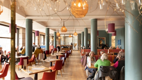 A bustling restaurant with two lines of green floor to ceiling pillars, red chairs and foliage among the ceiling hung lampshades.