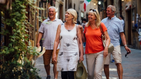 Two older white women in bright clothings walk smiling while two older white men walk behind them in a pedestrianised street. All are dressed in summer clothing.