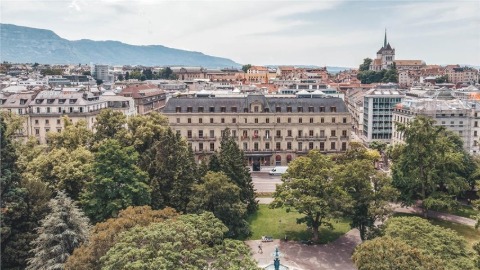 A view from a hill across the trees at the large neoclassical Metropole Geneva, the surrounding streets and mountains in the background.