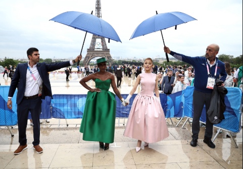 Cynthia Erivo and Ariana Grande in front of the Eiffel Tower with a security guard by each of they sides holding umbrellas above them