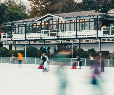 A white and brown two storey building overlooking a pop up ice rink, featuring blurred figures of ice skaters.