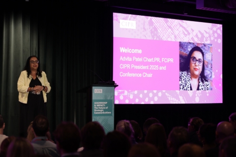 Advita Patel addressing the audience in the dark auditorium. To her right is a large pink screen with her name and photo.