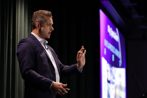 Jack Lewis on stage. In the background is the purple hue of his presentation