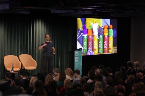 James Peach addressing the audience in the dark auditorium. To his right is a screen with brightly coloured smoothie bottles topped with miniature knitted hats.