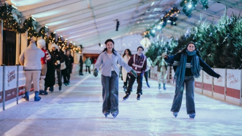 Three ice skaters skate towards the camera while others stand by the side railings. There are Christmas trees and decorations along each side and a white roof.