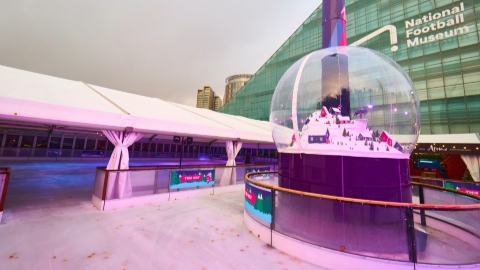 A pink hued colour photos of a covered ice rink and a large snow globe on a plinth in front of the triangular glass National Football Museum