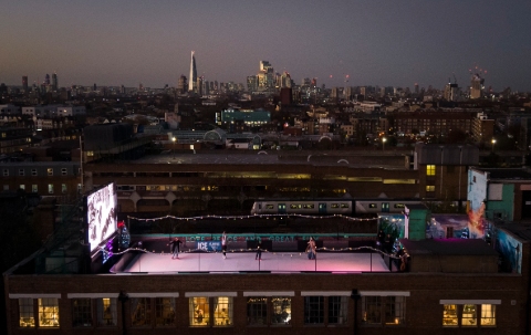 An aerial view of a rooftop ice rink, bathed in pink light on an otherwise dark night, with the London skyline in the distance 