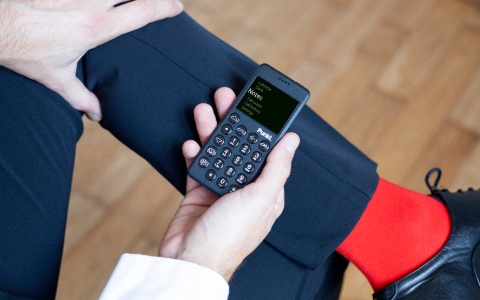 A male hand holds a Punkt mobile device on his crossed leg. He wears dark trousers, red sock and black shoes.