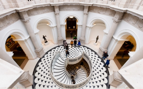 A bird's eye view of the rotunda at the Tate Britain featuring curved stone walls decorated with arches and a central spiral staircase patterned in black and white.