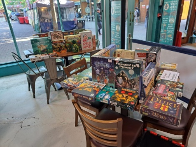Two tables in front of a floor to ceilings window that both have various board games stacked on top