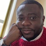 A portrait of Carl Kwaku Asiedu resting his cheek on his fist. Asiedu is a black man with short dark hair and bear who is wearing a red jumper.